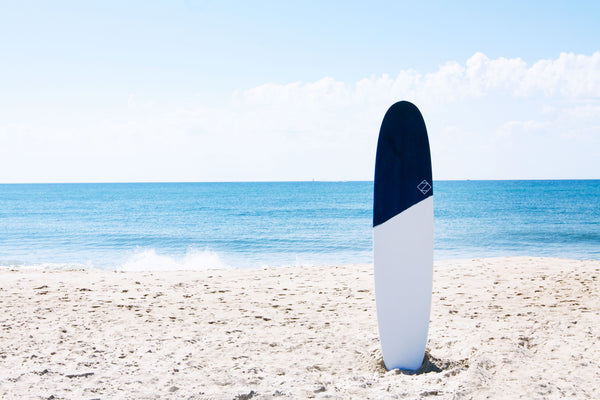 A single surfboard in dark and light blue stands tall on the beach amongst a picturesque view of the ocean