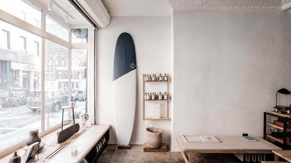A storefront in Soho with a tall surfboard in the center and a Frama shelving unit to its side. The window looks out to the streets of New York City