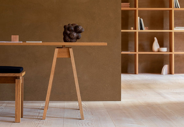 The clean lines of a desk and chair made of solid wood against an olive green wall. In the background shows a minimal bookcase with small objects