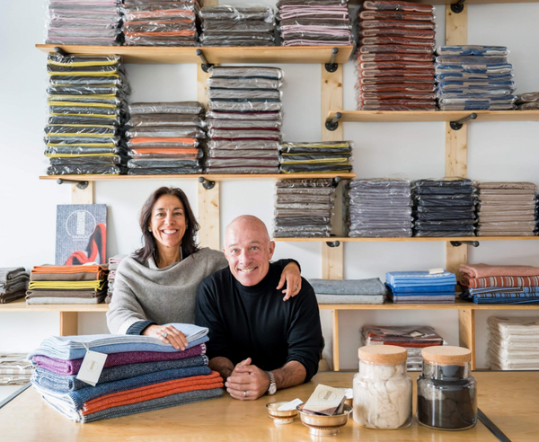 Bill and Betina Infante smiling in their office with Hangai Mountain Textiles throws stacked behind them on a wall